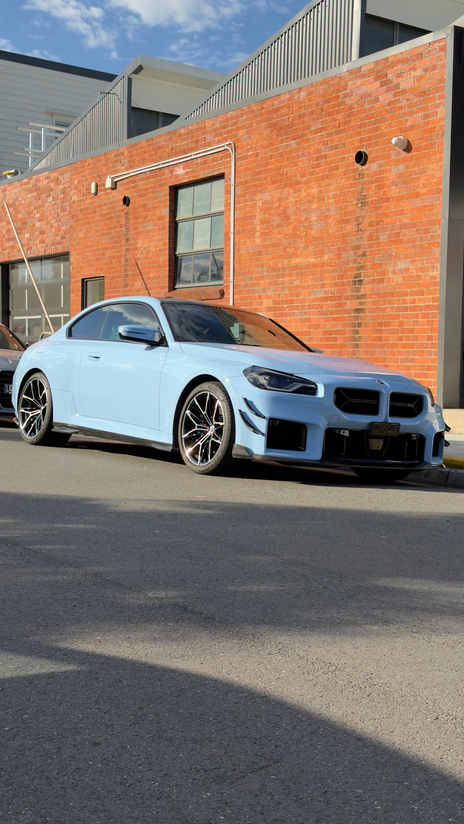 A light blue BMW M2 coupe parked on a street in front of a brick building under a blue sky with clouds.